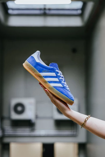 adidas Gazelle Indoor Blue. Clara hält den Schuh vor einem grauen Hintergrund in der Hand