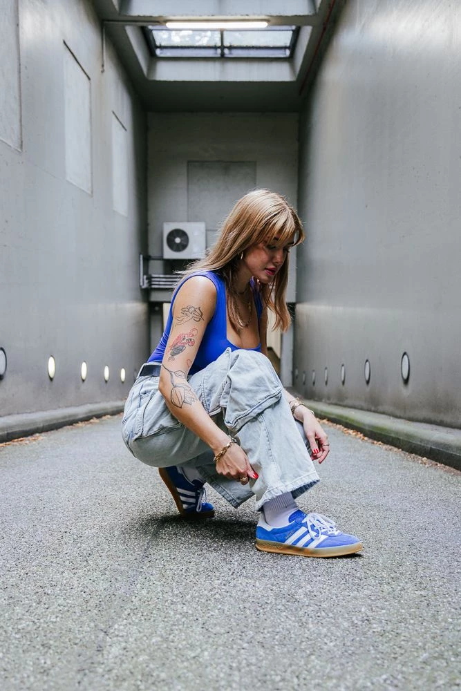 adidas Gazelle Indoor Blue Clara kniet vor einem grauen Hintergrund