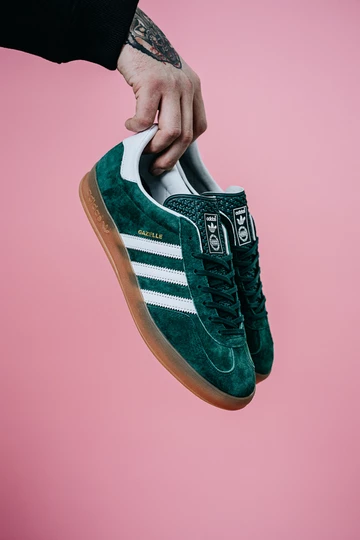 Christopher hält die adidas Gazelle Indoor Green auf rosafarbenem Rollhintergrund in der Hand