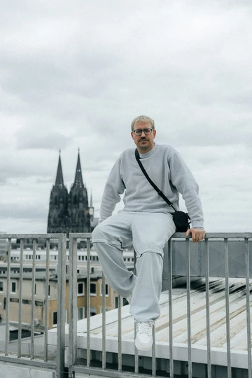 Christopher sitzt auf einer Brüstung, im Hintergrund ist der Kölner Dom und er trägt den BSTN x Real Madrid x adidas Rivalry Low 86