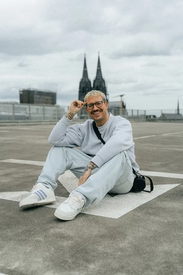 Christopher sitzt auf einem Parkplatz, im Hintergrund ist der Kölner Dom und er trägt den BSTN x Real Madrid x adidas Rivalry Low 86