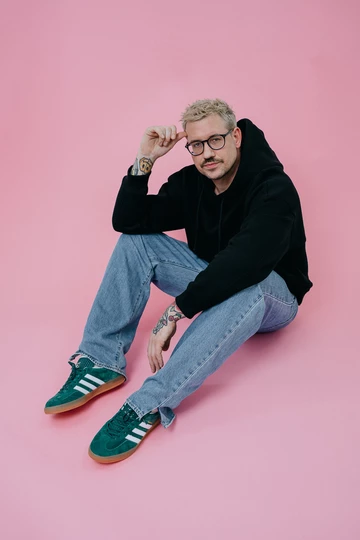 Christopher sitzt auf einem rosafarbenem Rollhintergrund und trägt die adidas Gazelle Indoor Green am Fuß