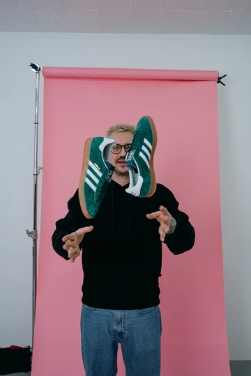 Christopher steht vor einem rosafarbenem Rollhintergrind und wirft die adidas Gazelle Indoor Green hoch