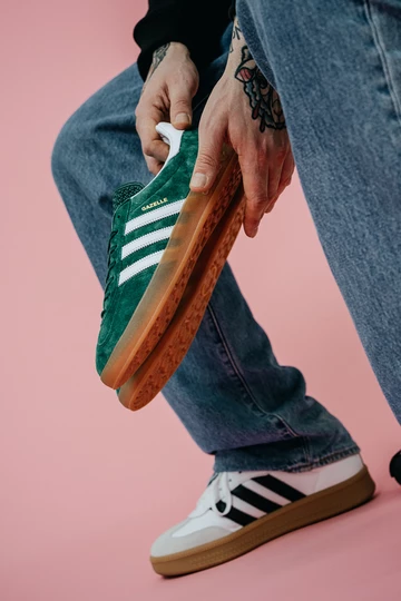 Christopher hält die adidas Gazelle Indoor Green auf rosafarbenem Rollhintergrund in der Hand