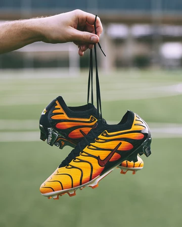 Jeman hält den Air Max Plus Mercurial Vapor 15 Pimento auf einem Fußballplatz in der Hand